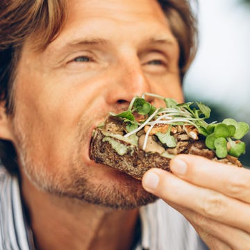 man with medium length hair and a beard enjoys eating an avocado toast with mushrooms and microgreens. his eyes are closed, savoring the taste.